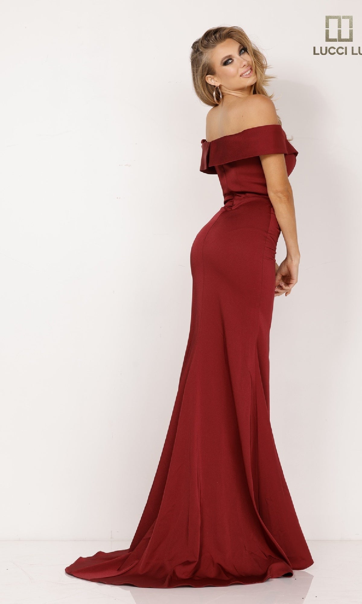 A woman poses against a white background, smiling over her shoulder in the Lucci Lu 8262 burgundy off-the-shoulder crepe prom dress. The Lucci Lu logo is displayed in the top right corner.